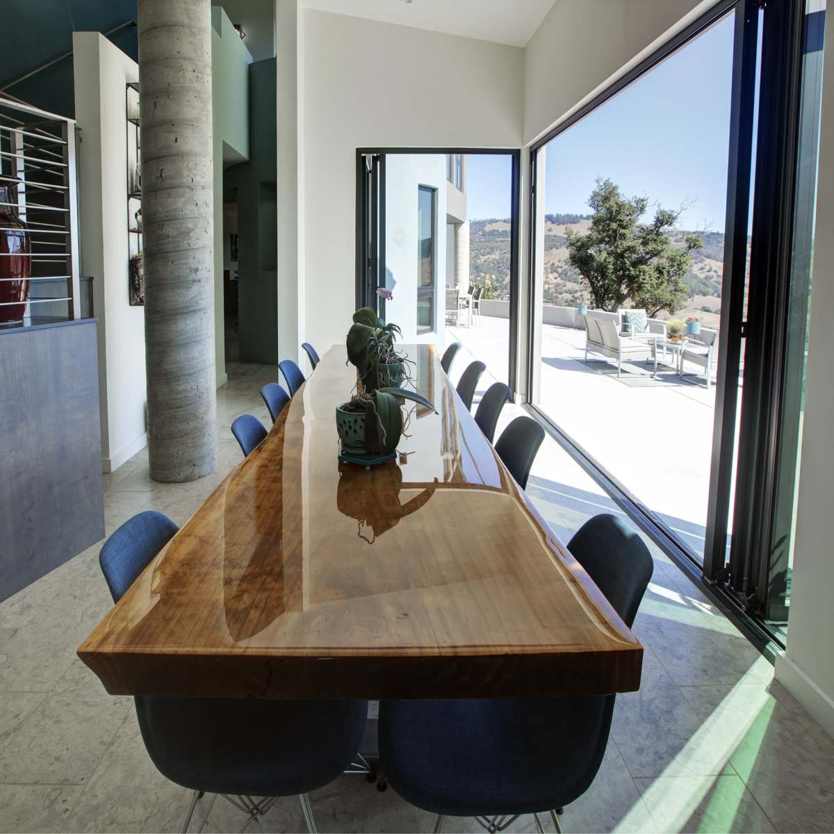A long wooden dining table with blue chairs is positioned in a modern interior space featuring large windows that open to a view outside.