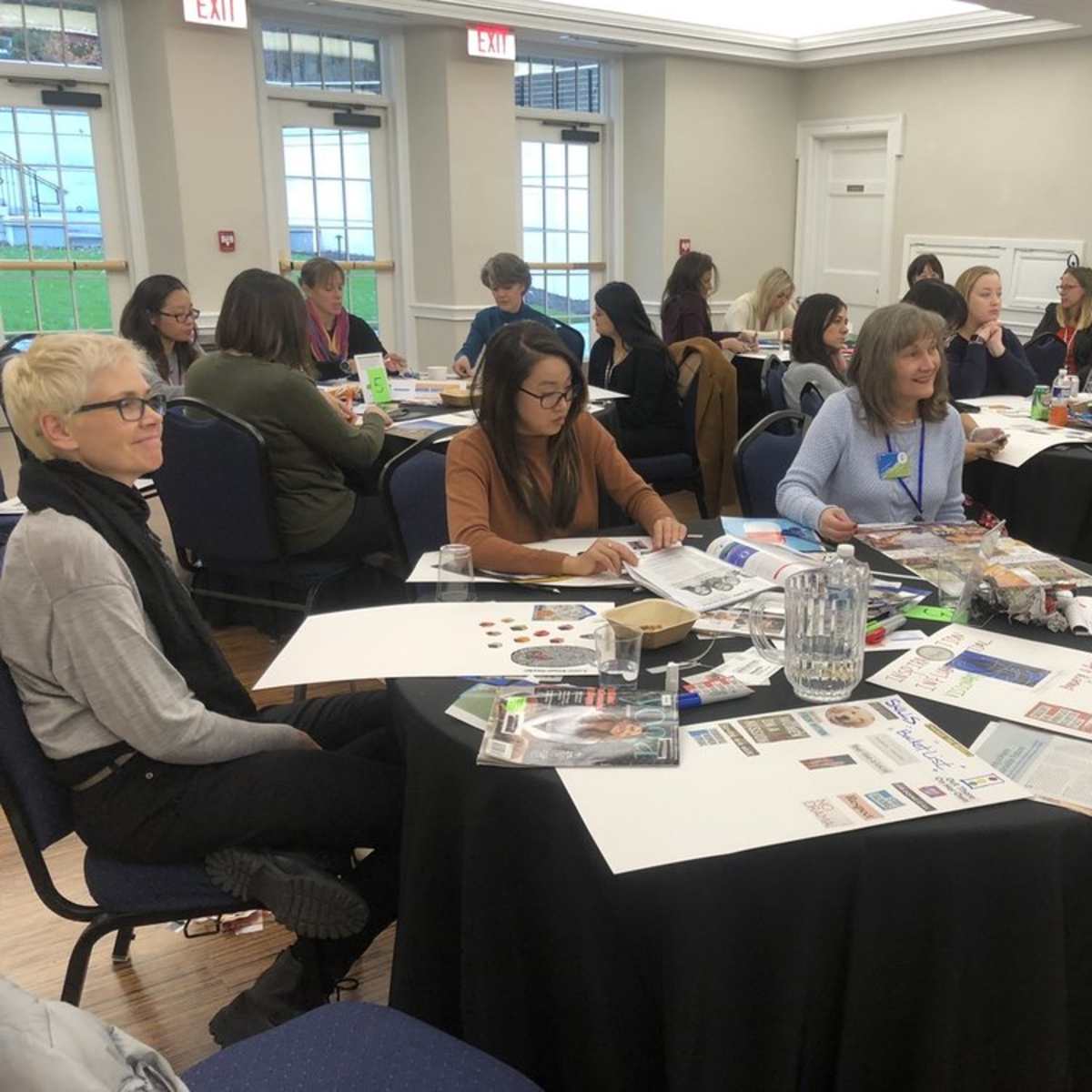 A group of people sits around tables covered with papers, magazines, and craft supplies during a workshop or meeting in a well-lit room.