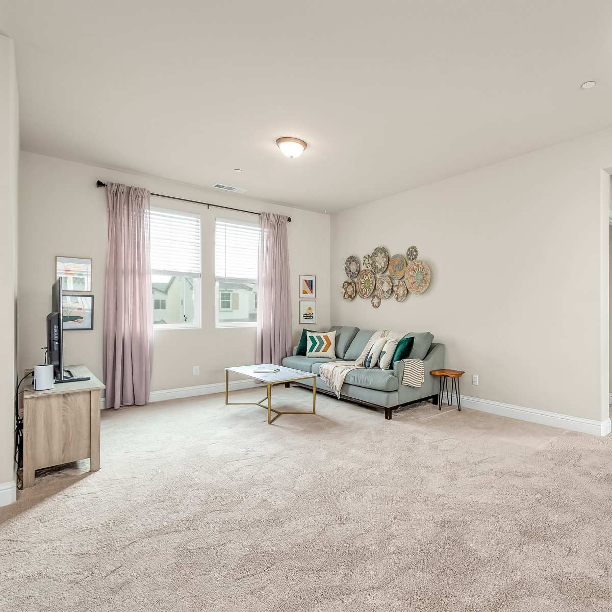 A spacious living room with a light carpet, a sofa adorned with colorful cushions, a coffee table, and wall decor, complemented by natural light from the two windows.