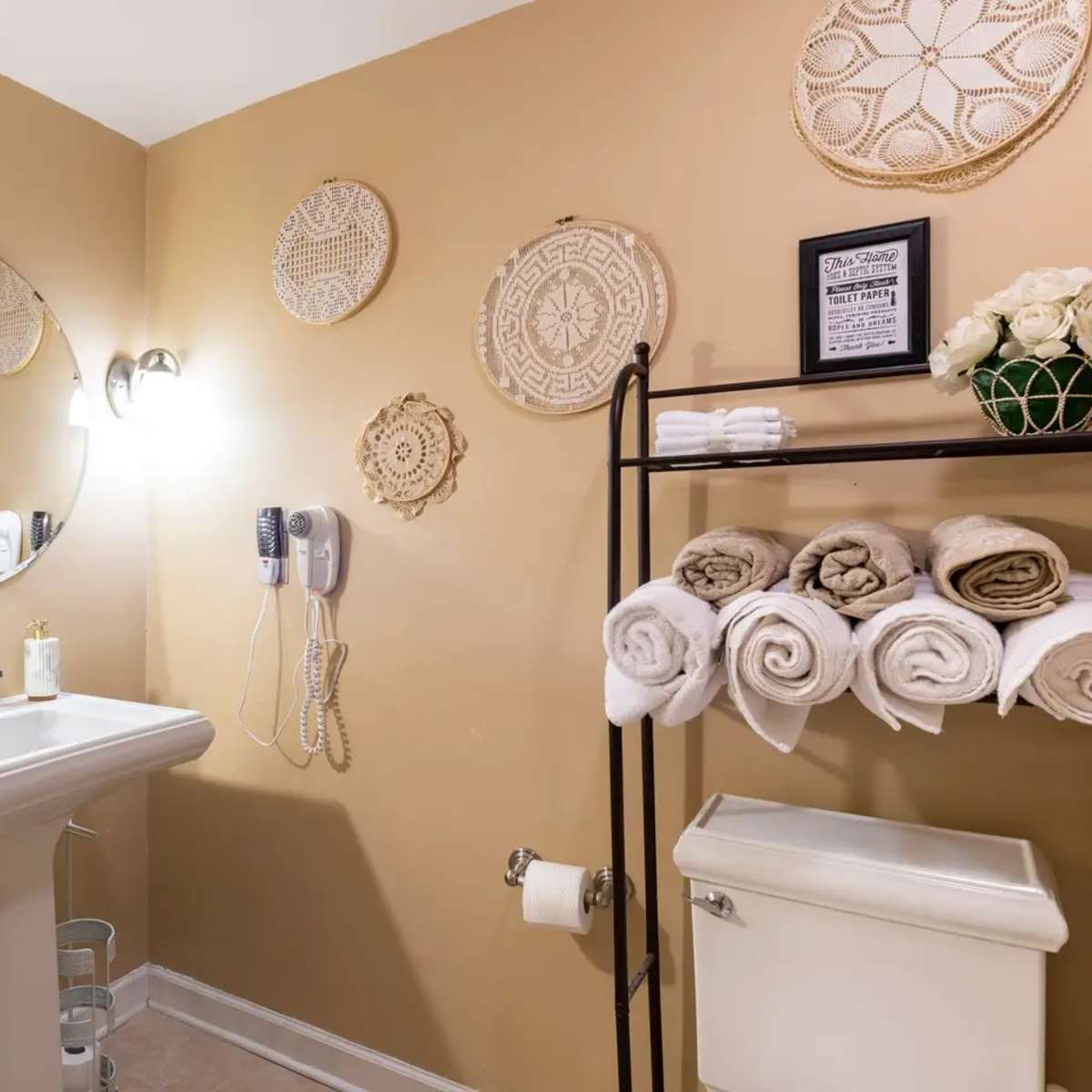 A bathroom with a round mirror above a white sink, beige walls adorned with decorative wall hangings, and neatly arranged folded towels on a rack.