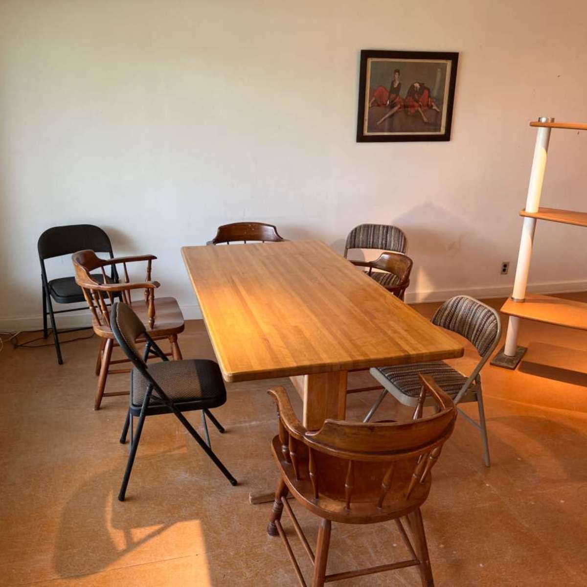 A wooden table surrounded by six chairs sits in an empty room with natural light streaming through a window.