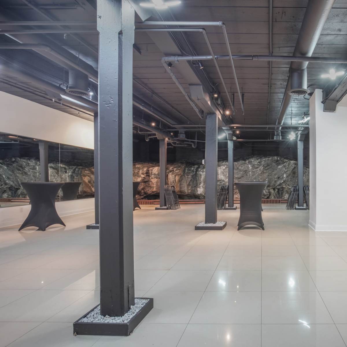 An empty event space with polished white tiles, black columns, and two high tables arranged in the center.
