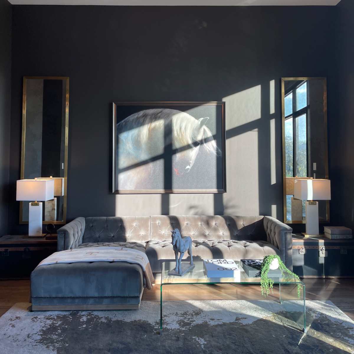 The living room features a sectional sofa, a large abstract painting, floor-to-ceiling windows, and a sunburst chandelier, with natural light casting shadows on the furniture.