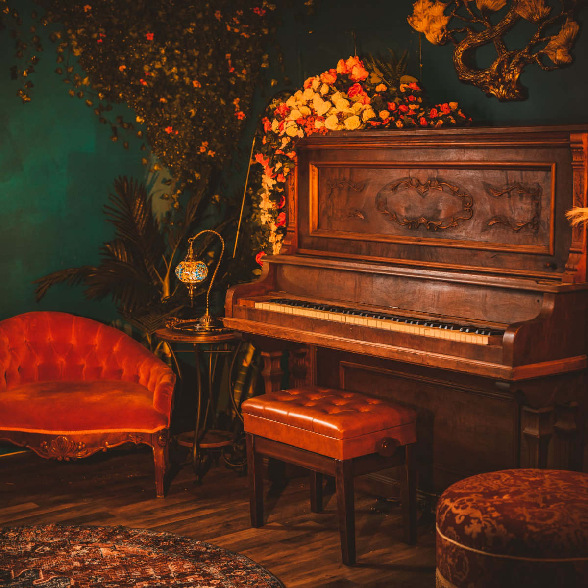 A vintage wooden piano sits next to an ornate orange couch in a warmly decorated room filled with flowers and soft lighting.