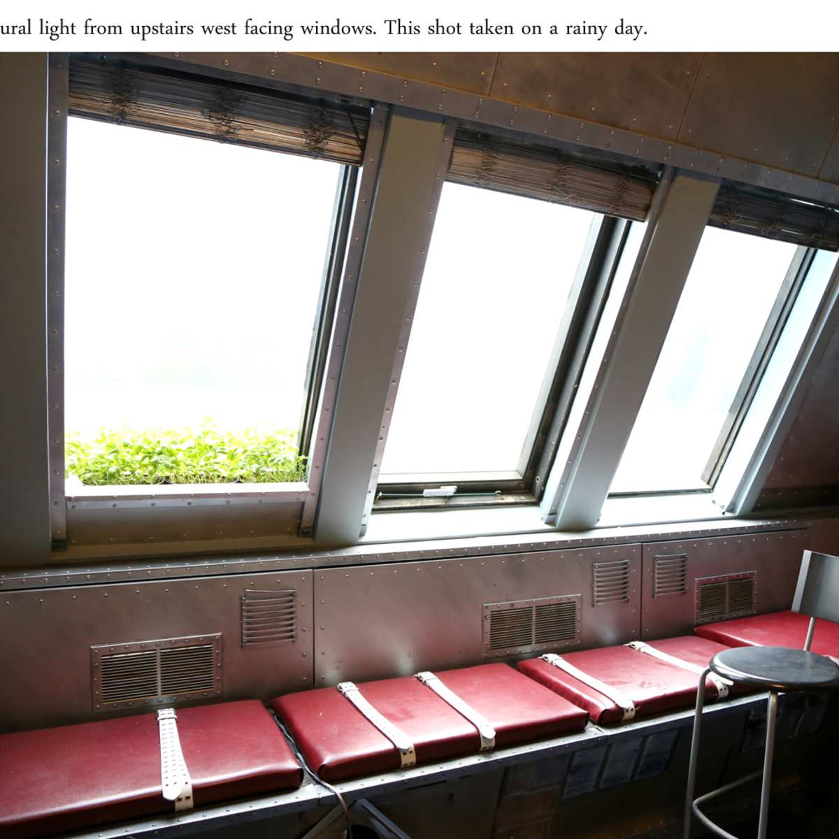 A seating area with red chairs and a barstool next to large, west-facing windows that let in natural light on a rainy day.