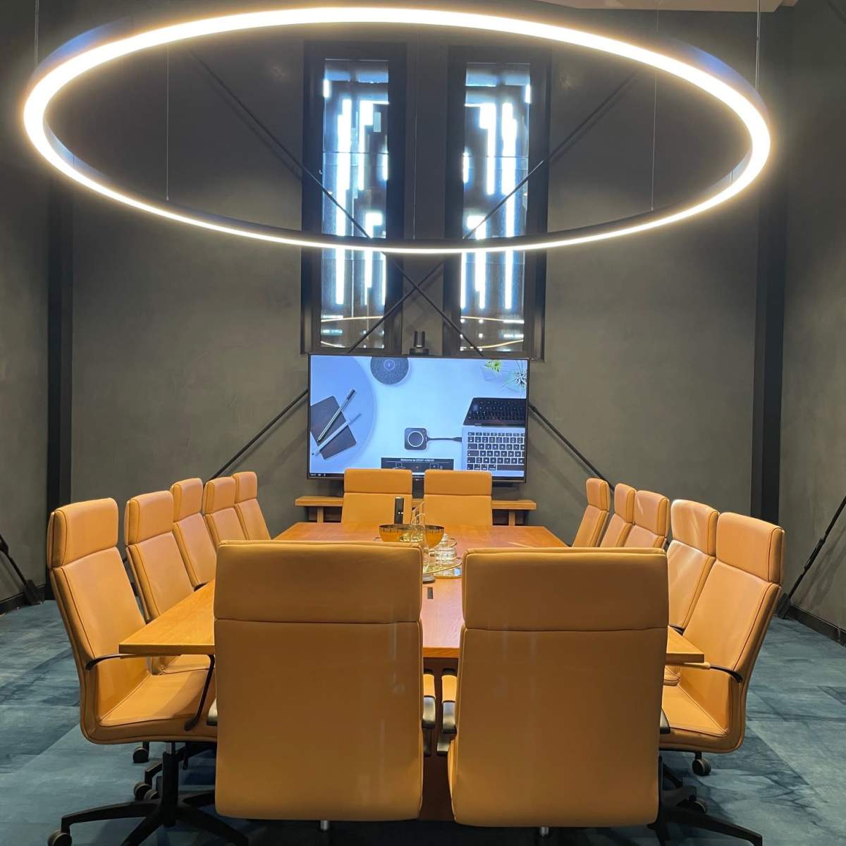 A modern conference room featuring a large wooden table surrounded by orange chairs and a circular light fixture overhead, with a screen displaying a laptop and office-related items.