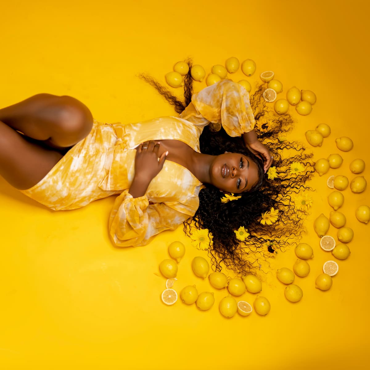 A person in a yellow outfit lies on a yellow floor surrounded by lemons and flower petals.