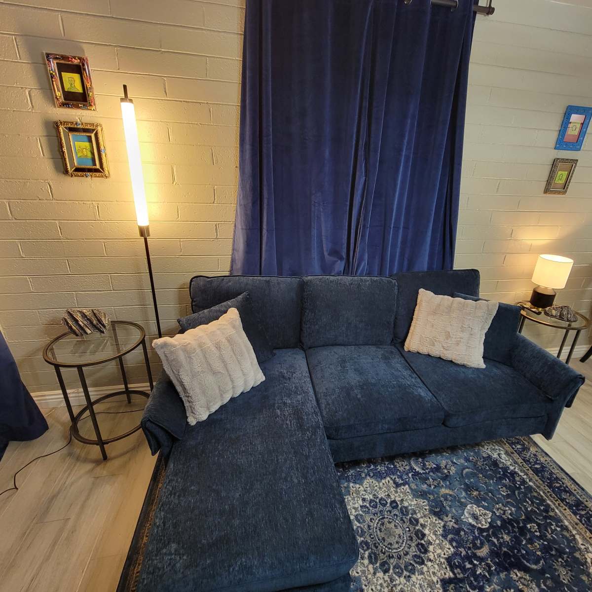 A living room with a dark blue sectional sofa, a standing lamp, and a round table with a yellow chair, set against a backdrop of blue curtains and framed artwork on the walls.