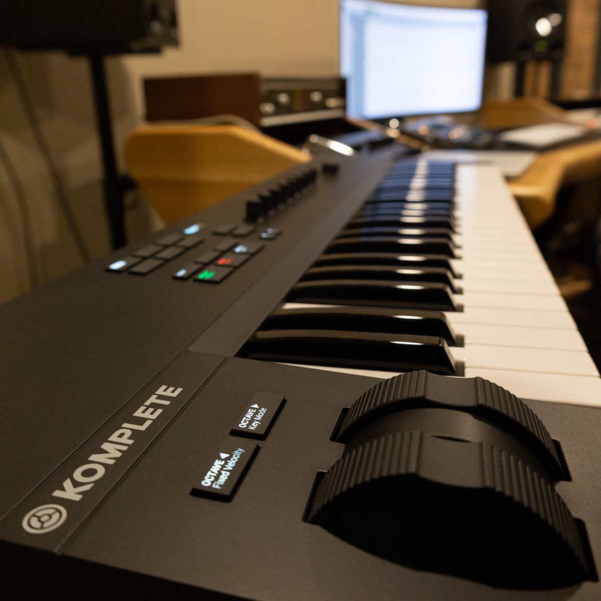A close-up of a music keyboard controller with a computer setup and studio monitors in the background.