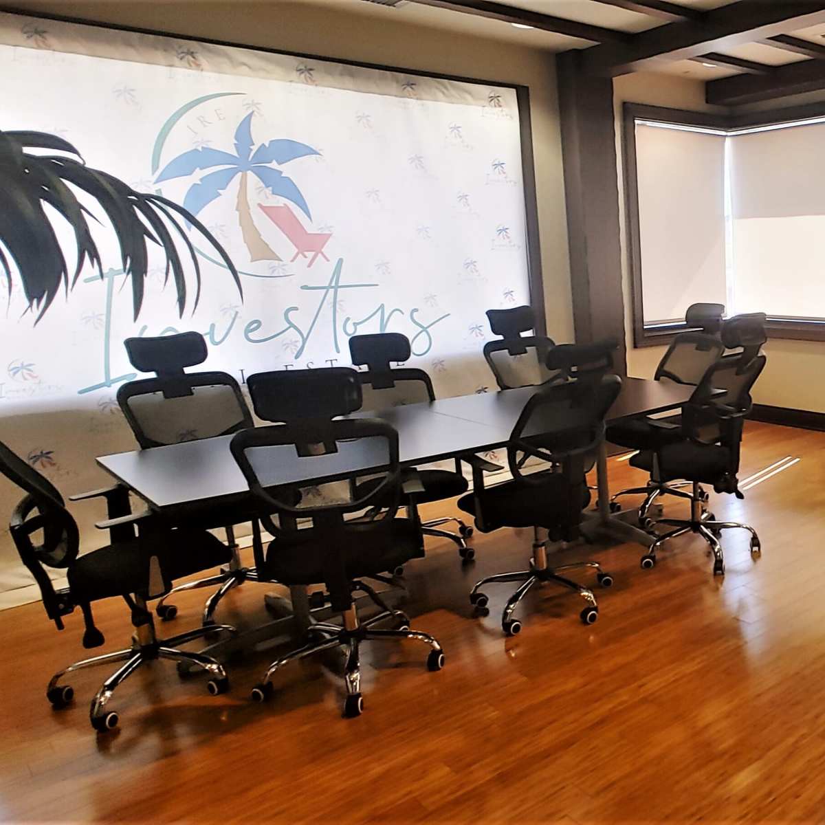 A conference room features a large table with black chairs, a round bar table with stools, a beige lounge couch, and a backdrop displaying a logo with palm trees.