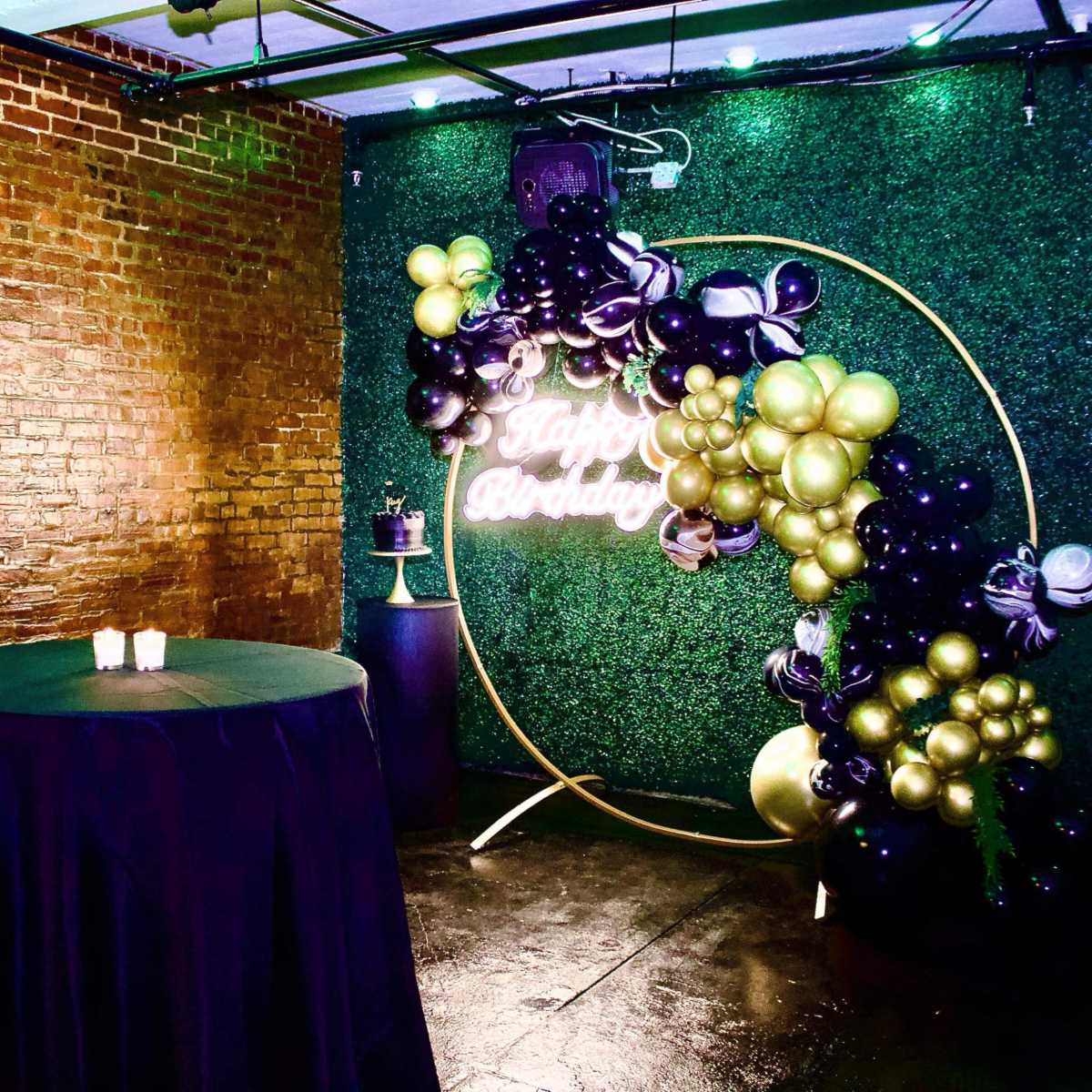 A decorated event space with a green wall, a circular balloon arch, and a table covered in black fabric.