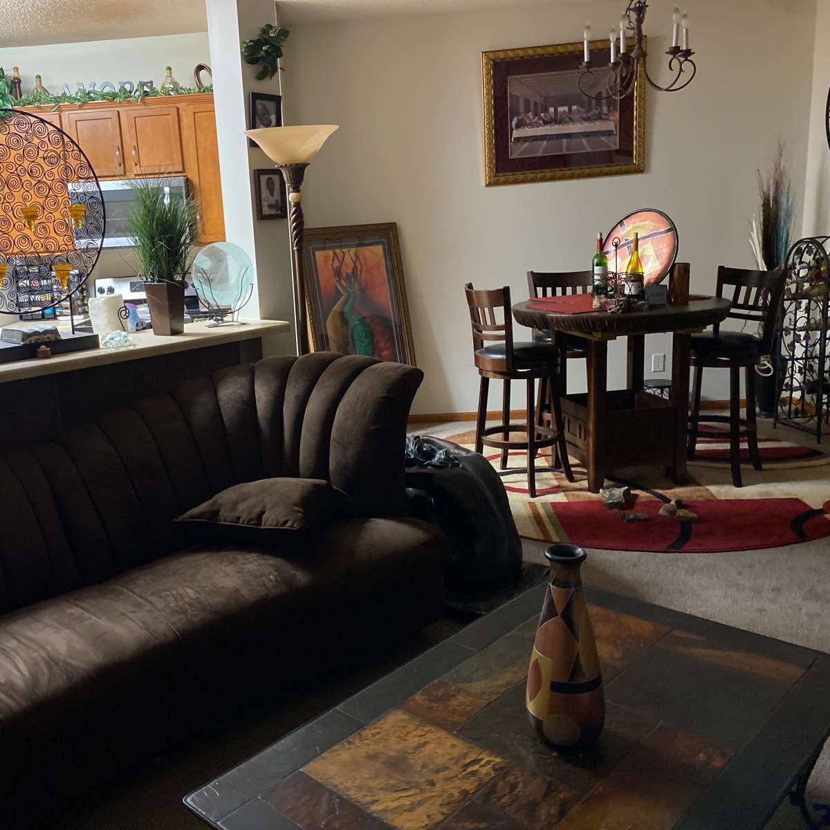 A living room with a brown sofa, a coffee table, a dining area with a small table and chairs, and a kitchen visible in the background.