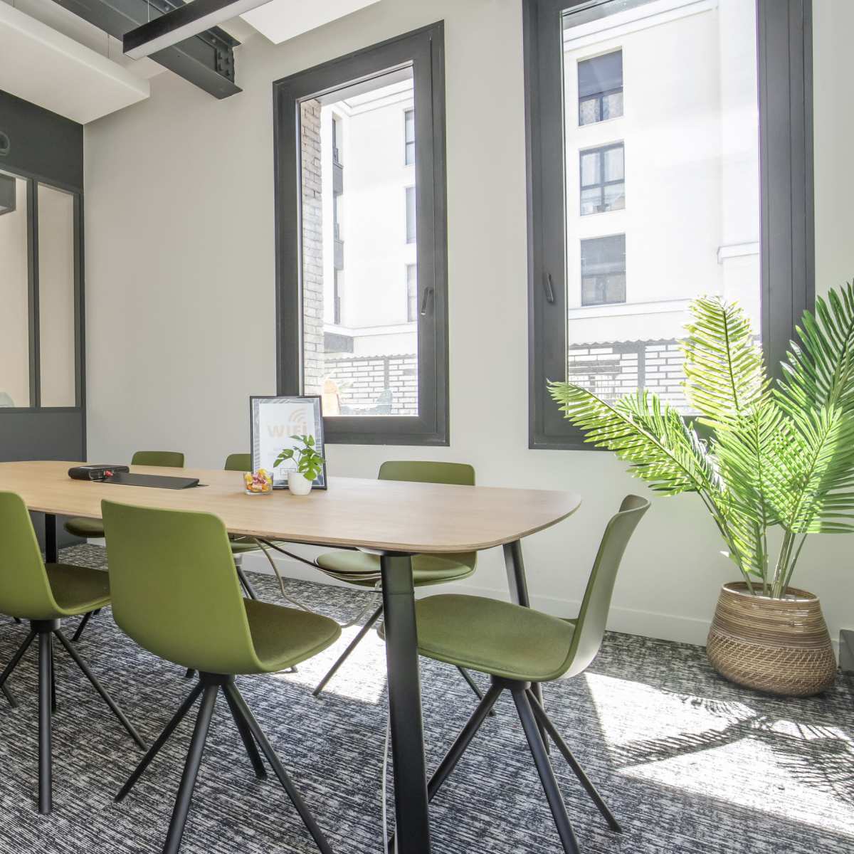 A modern conference room features a long wooden table surrounded by green chairs, with large windows allowing natural light to illuminate the space.