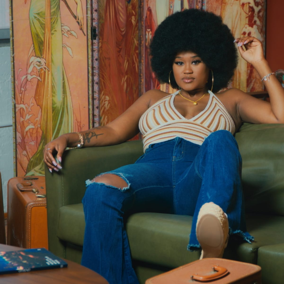 A woman with an afro hairstyle sits casually on a green couch, wearing a striped top and ripped jeans, surrounded by colorful decor and vintage items.