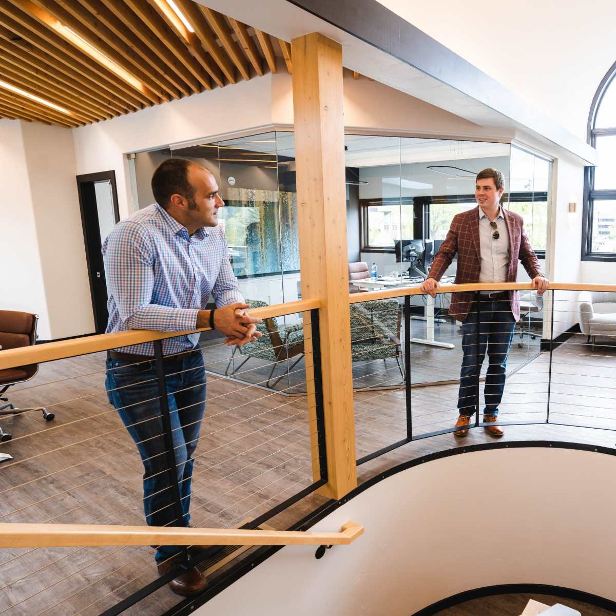 Two men converse while leaning on a wooden railing above a circular staircase in a modern office space, with a glass-walled meeting area in the background.