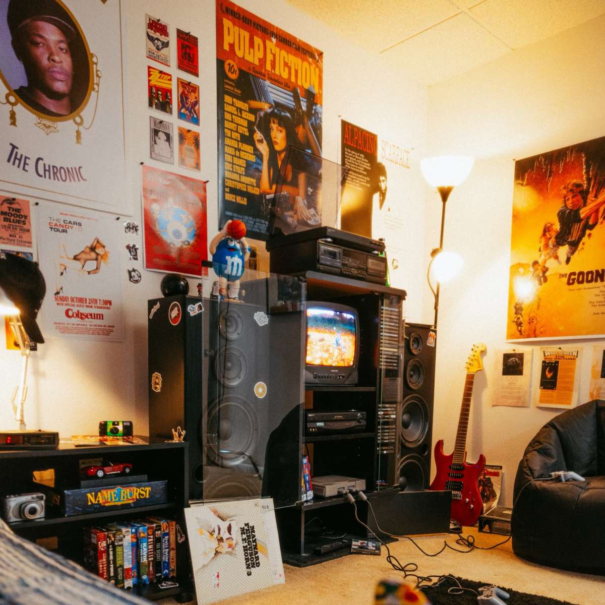 A retro-styled living room features a vintage television, music posters on the walls, a guitar, and a skateboard resting on a colorful blanket.