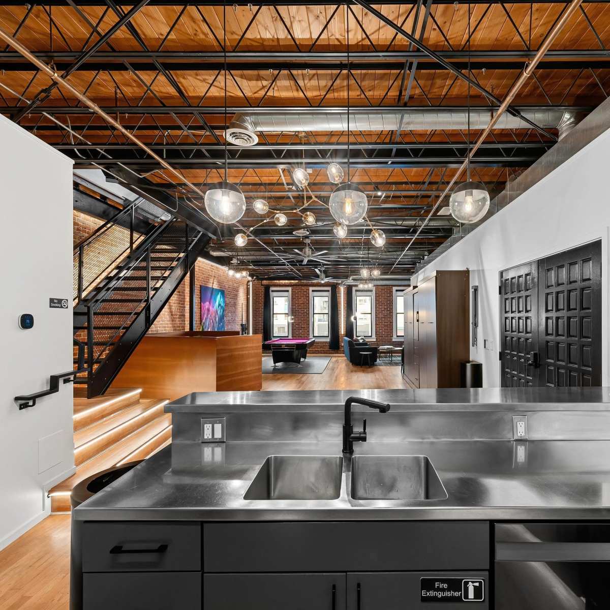 A modern kitchen area with stainless steel fixtures, leading into a spacious open-concept living space featuring wooden floors and industrial-style lighting.