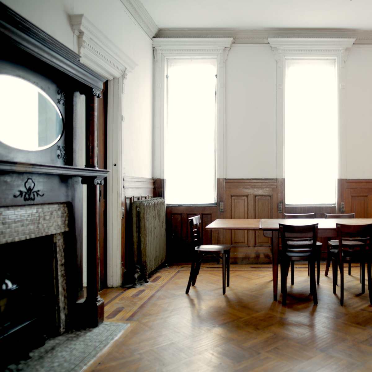 A large, empty room features a wooden table surrounded by chairs, with tall windows allowing natural light to enter.