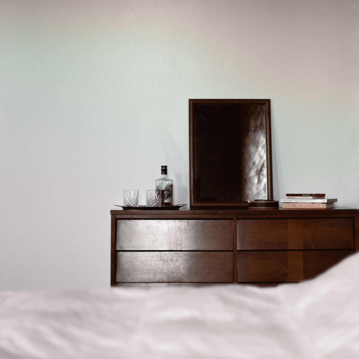 A wooden dresser with a mirror sits against a wall, accompanied by a bottle and glassware on top.