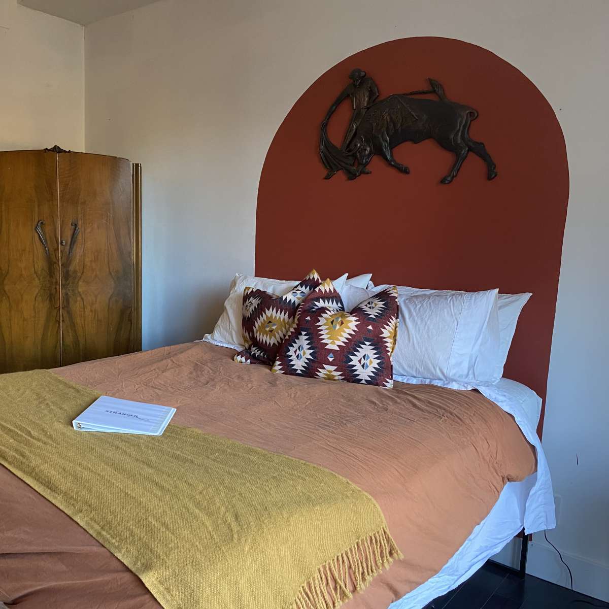 A cozy bedroom featuring a large bed with a brown spread and patterned pillows, a wooden wardrobe, a framed cowboy-themed artwork on the wall, and a distinctive red arch with a bull sculpture above the headboard.