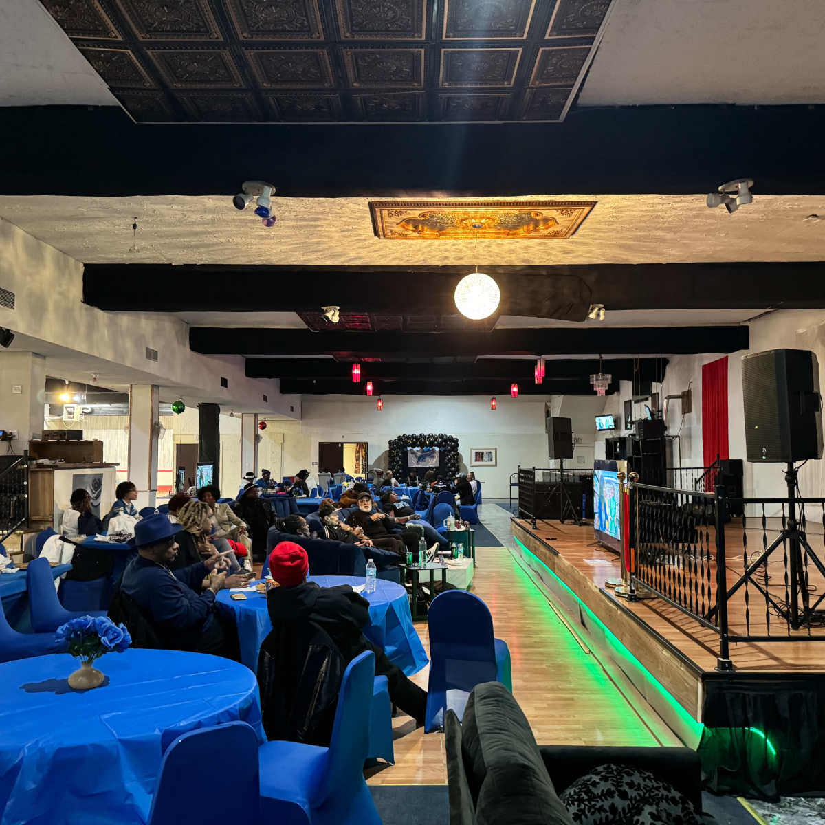 A spacious indoor venue with blue tables and chairs, where a group of people are seated, facing a stage with a microphone and a screen.
