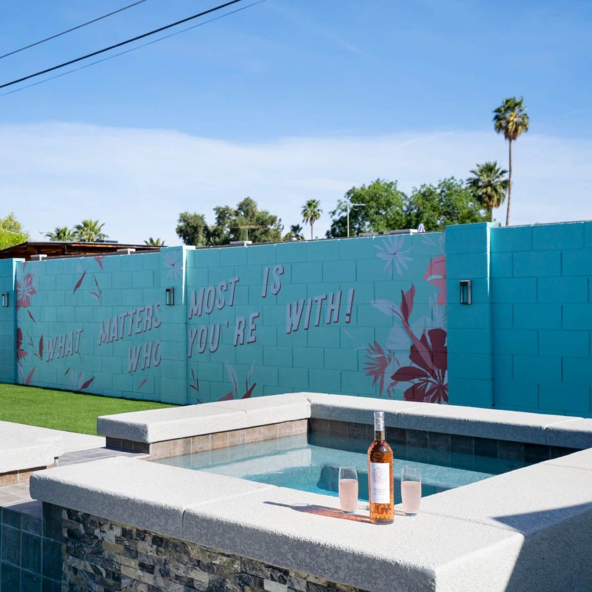 A hot tub sits beside a swimming pool, with a wall behind it featuring the phrase "WHAT MATTERS MOST IS WHO YOU'RE WITH" in stylized lettering, surrounded by palm trees and blue skies.