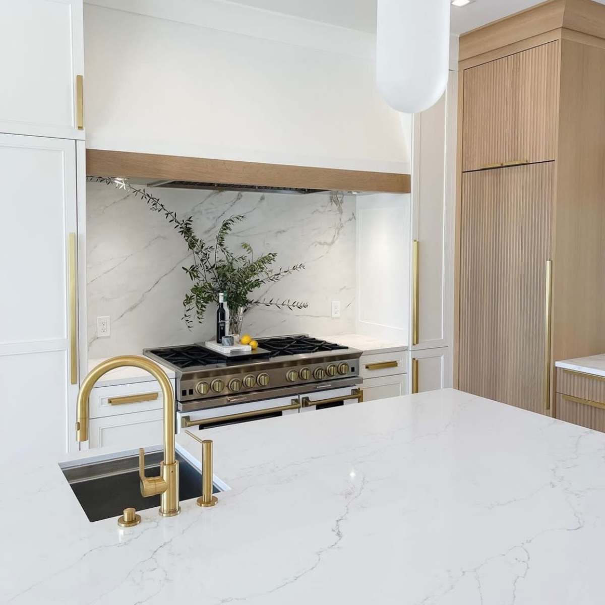 A modern kitchen features a marble backsplash, stainless steel stove, and a large white island with a gold faucet.