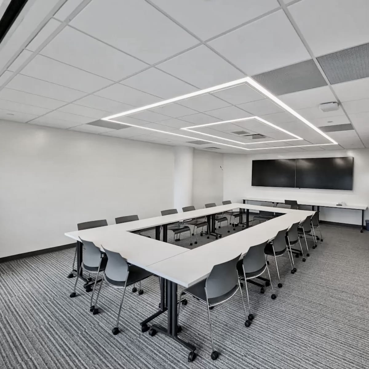 A conference room features a rectangular table surrounded by chairs, with a large screen mounted on the wall.