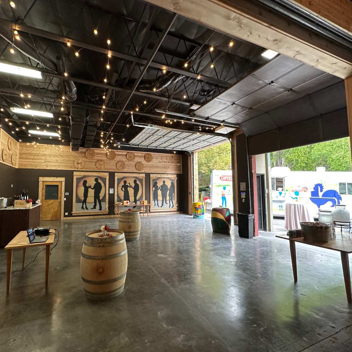 An indoor event space features wooden walls, silhouetted figures, wine barrels, and string lights.