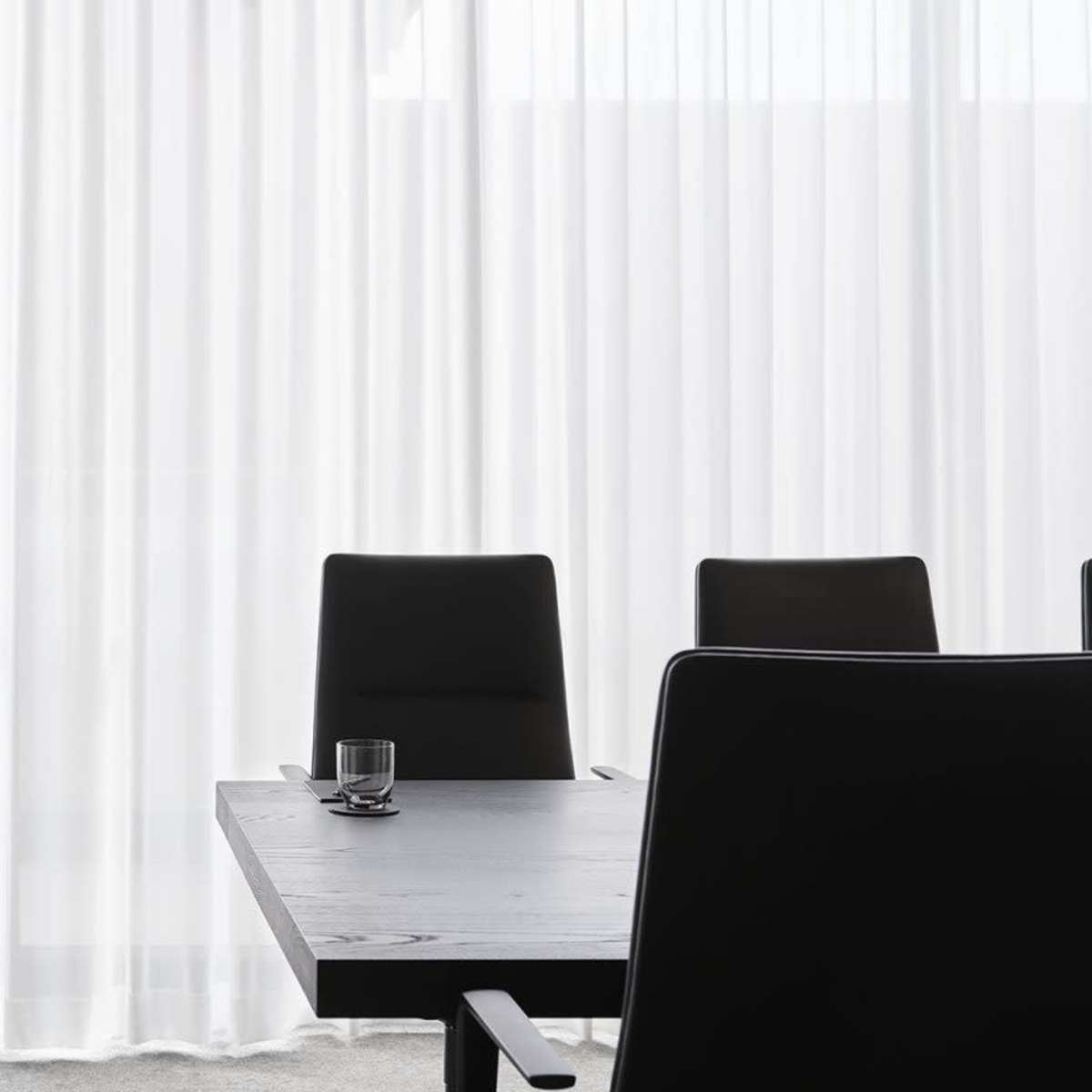 A modern conference room features a large table and black chairs, with a cart displaying glasses and bottles against a backdrop of sheer white curtains.