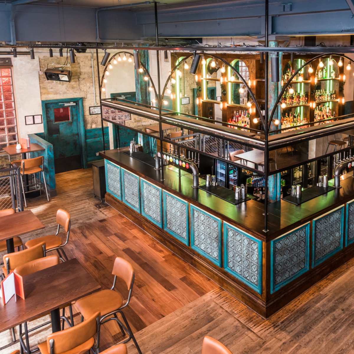 An elevated view of a modern bar area with a glass partition, illuminated shelves, and wooden flooring in a lively restaurant setting.