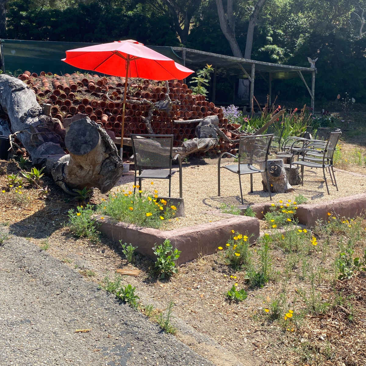A red umbrella shades a circular seating area surrounded by potted plants and gravel.