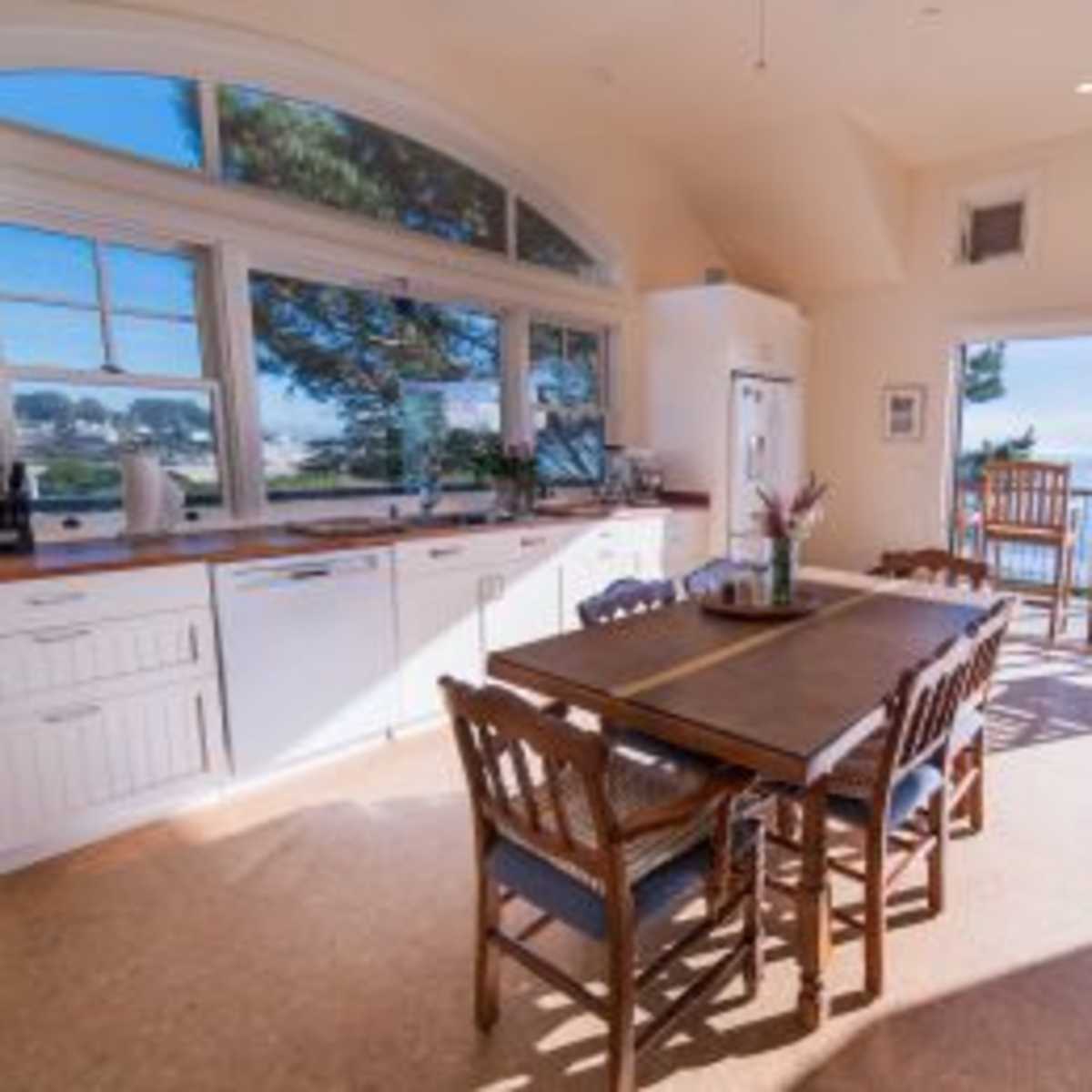 A bright kitchen and dining area featuring a large wooden table surrounded by chairs, with a view of the outdoors visible through large windows.