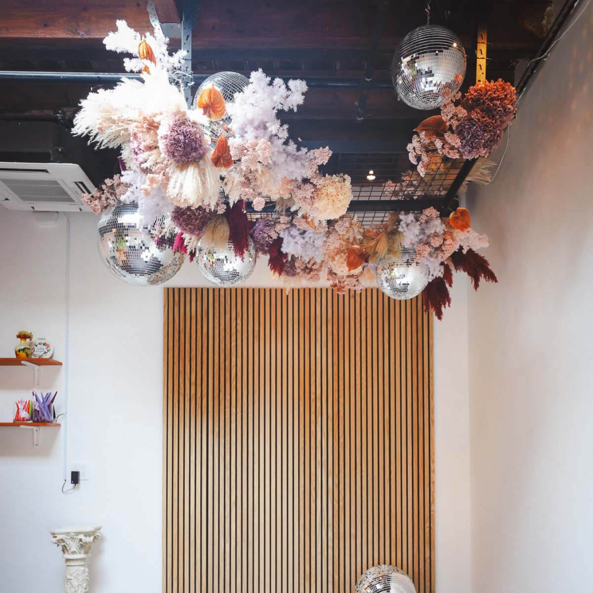 A room with six white chairs arranged in front of a wooden wall, decorated with colorful floral installations and hanging disco balls.