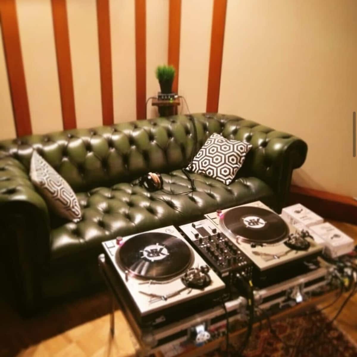 A green leather chesterfield sofa next to a pair of turntables set on a glass table in a room with wooden wall panels.