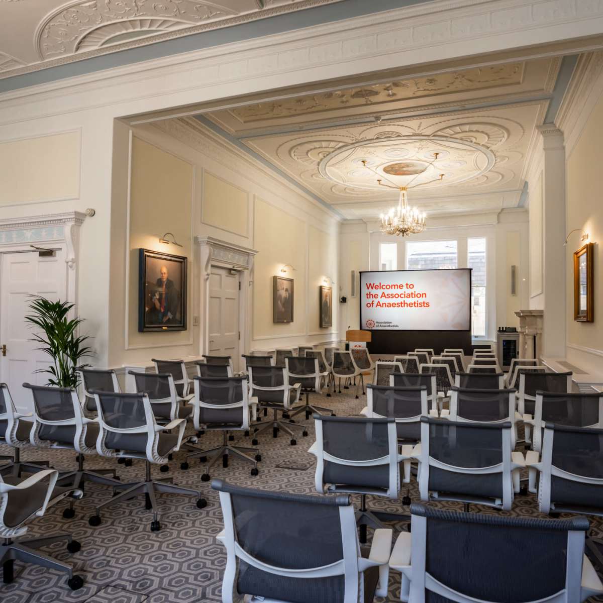 A spacious meeting room with rows of empty chairs facing a presentation screen displaying "Welcome to the Association of Anaesthetists."