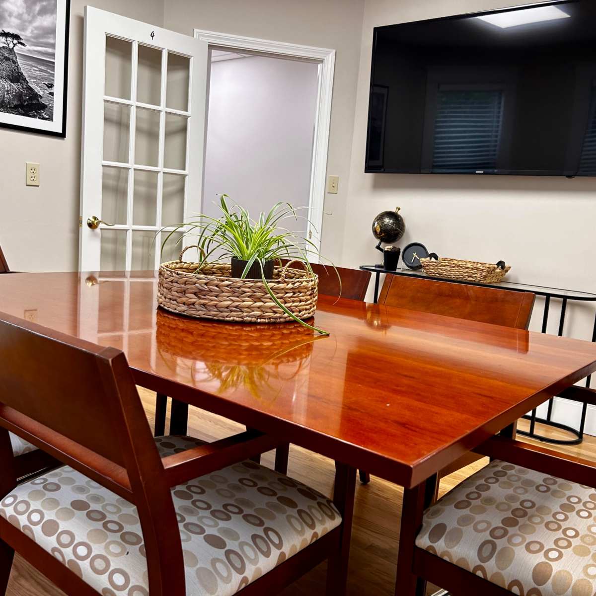 A dining area features a wooden table surrounded by upholstered chairs, with a decorative plant in a basket at the center and a wall-mounted television in the background.