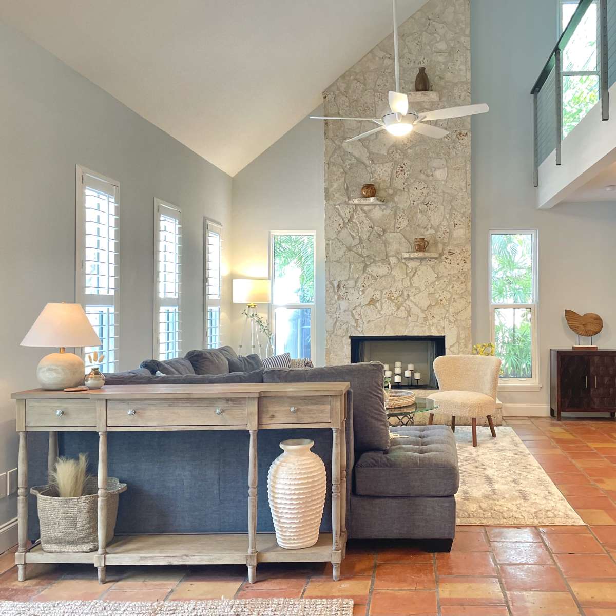 A spacious living room features a gray sofa, a wooden console table, and a stone fireplace with a tall ceiling, illuminated by natural light.