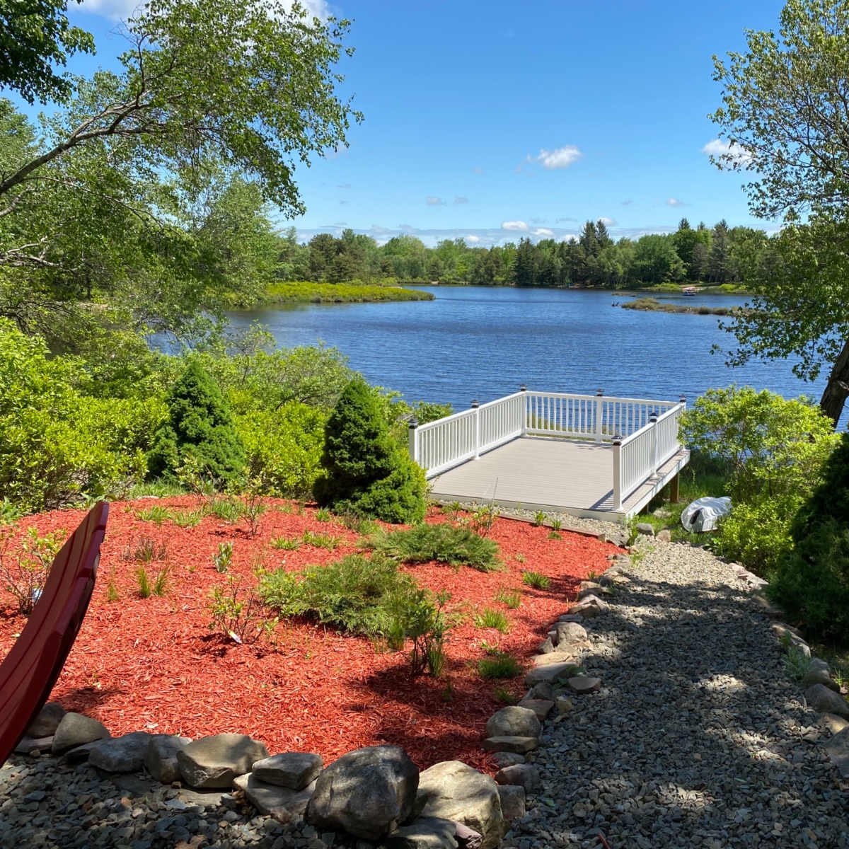 The scene features a landscaped area with a gravel path leading to a wooden deck overlooking a calm lake surrounded by trees and greenery.