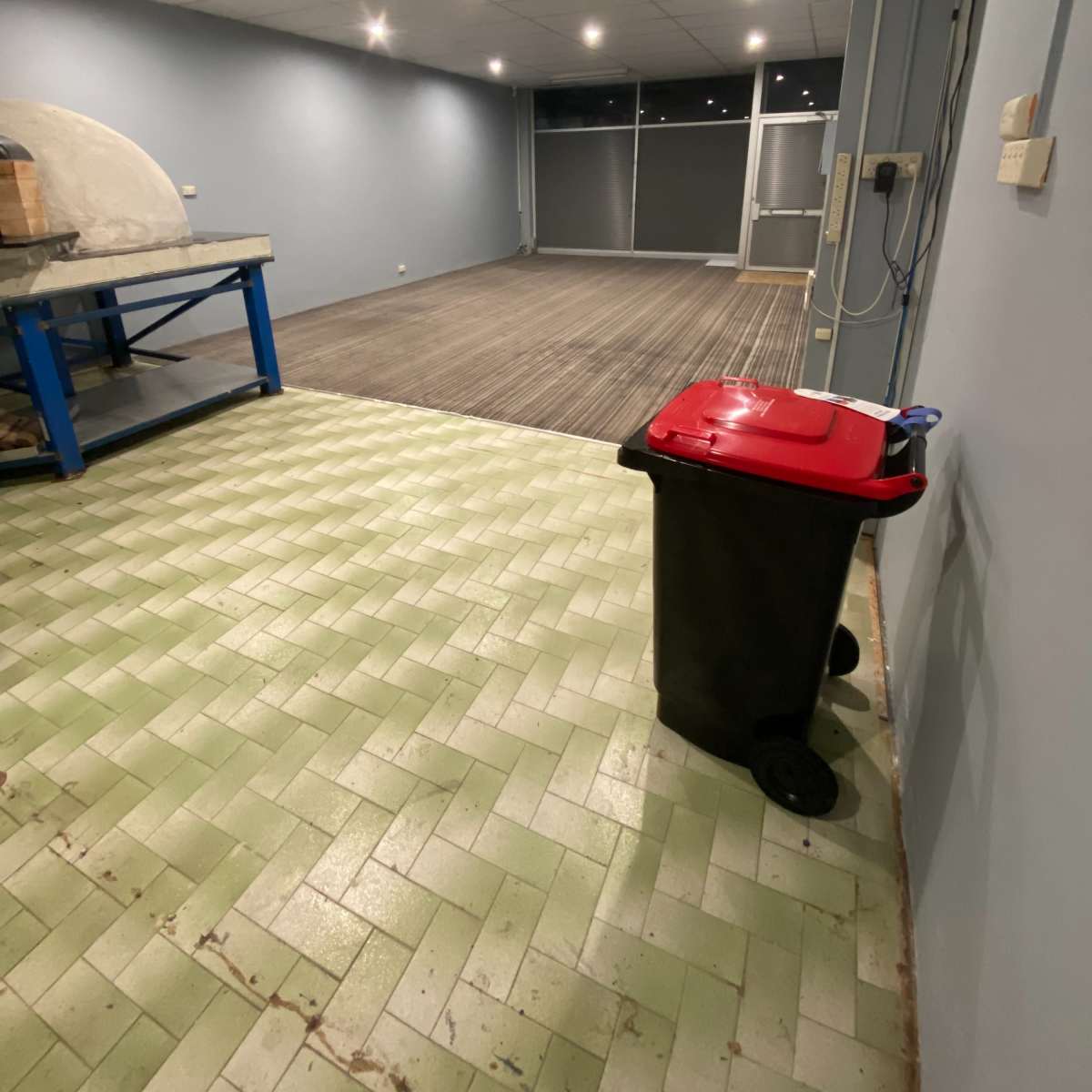 An empty room with light green and gray tiled flooring, a red-lidded trash bin, and blue tables along one wall.