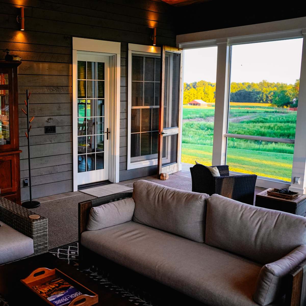 A cozy porch area with a wooden cabinet, two seating arrangements, and large windows overlooking a green field.
