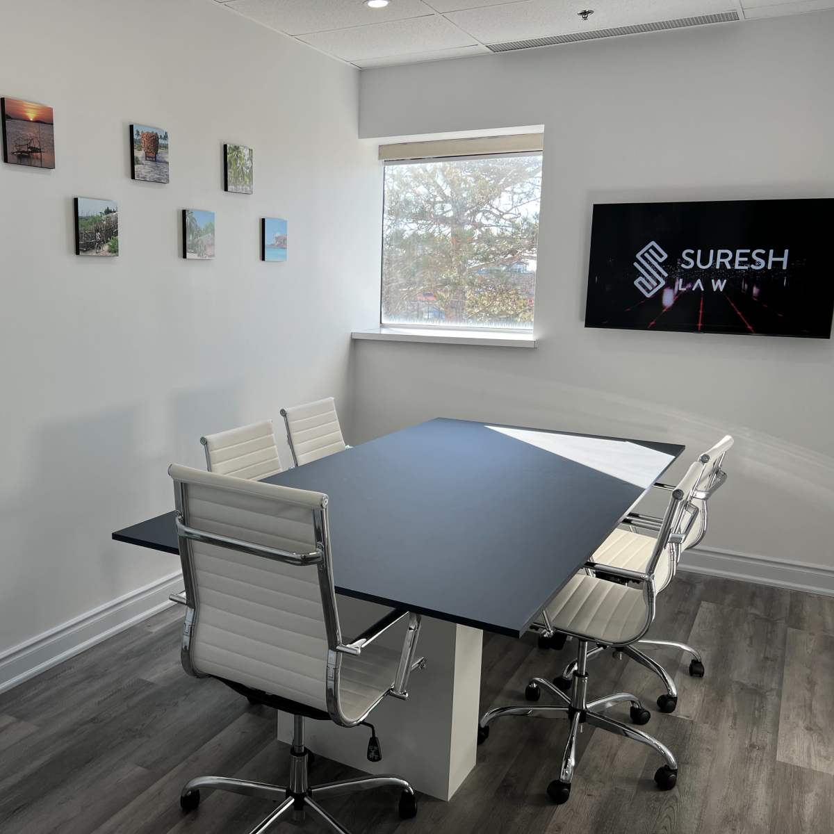 A modern conference room features a long black table surrounded by white chairs, with paintings on the wall and a television displaying "SURESH LAW."