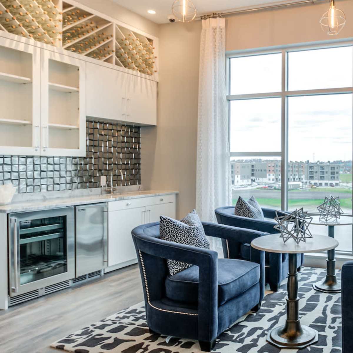 A stylish lounge area with four blue velvet chairs arranged around a small round table, large windows providing a view of the outdoors, and a modern kitchen in the background.