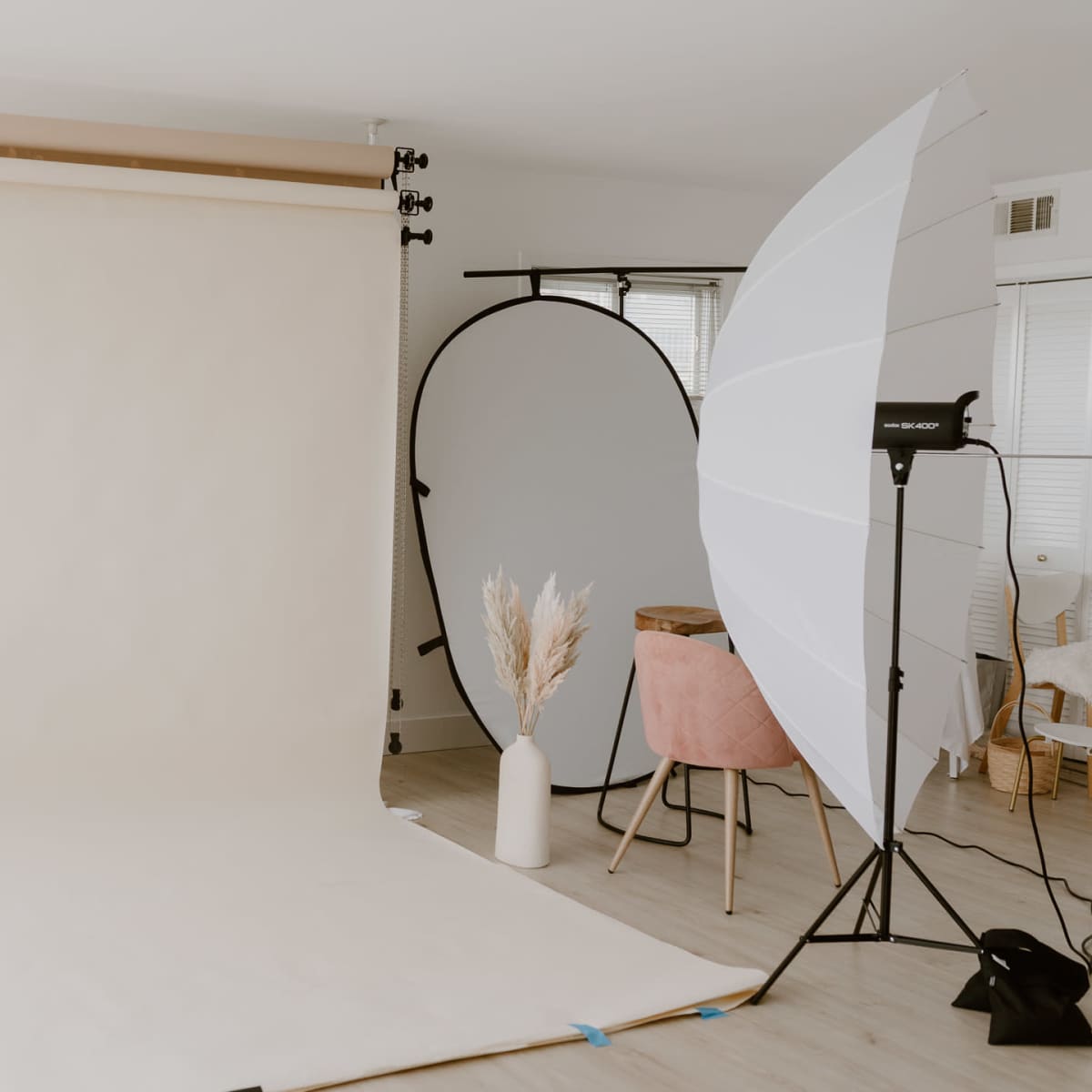 A photography studio setup with a soft beige backdrop, a round mirror, a large softbox light, a pink chair, and a vase with dried plants.