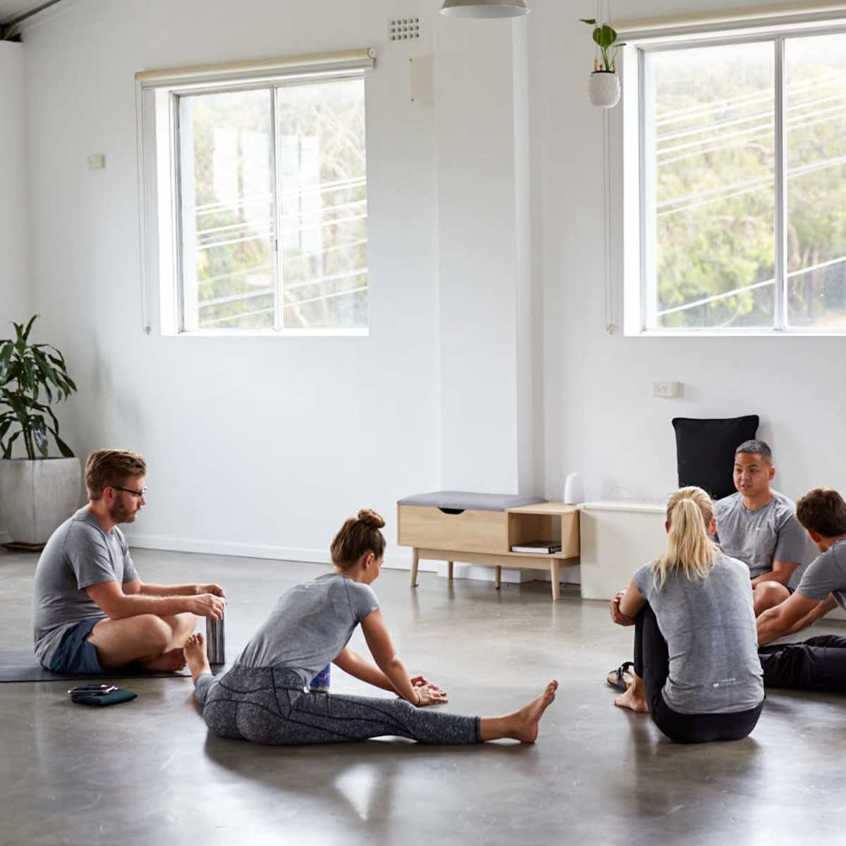 A group of five people sits on the floor in a light-filled room, engaging in conversation while some stretch on yoga mats.