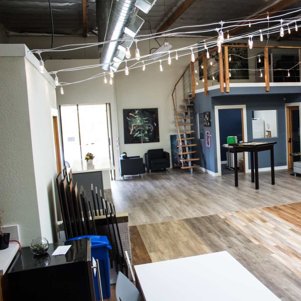 An open office space featuring wooden floors, modern furniture, and a staircase leading to a loft area, with string lights overhead.