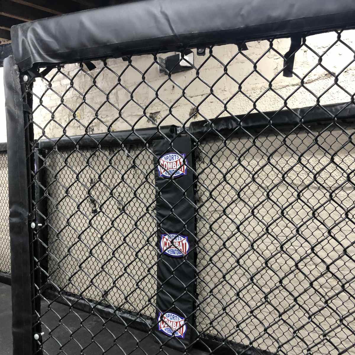 A section of a padded fighting cage with a black mesh fence and a logo for "Sport Combat" on the padding.