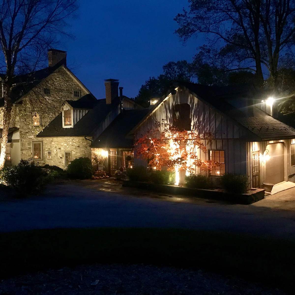 A stone and wood house is illuminated by lights at dusk, with trees and shrubs in the foreground.