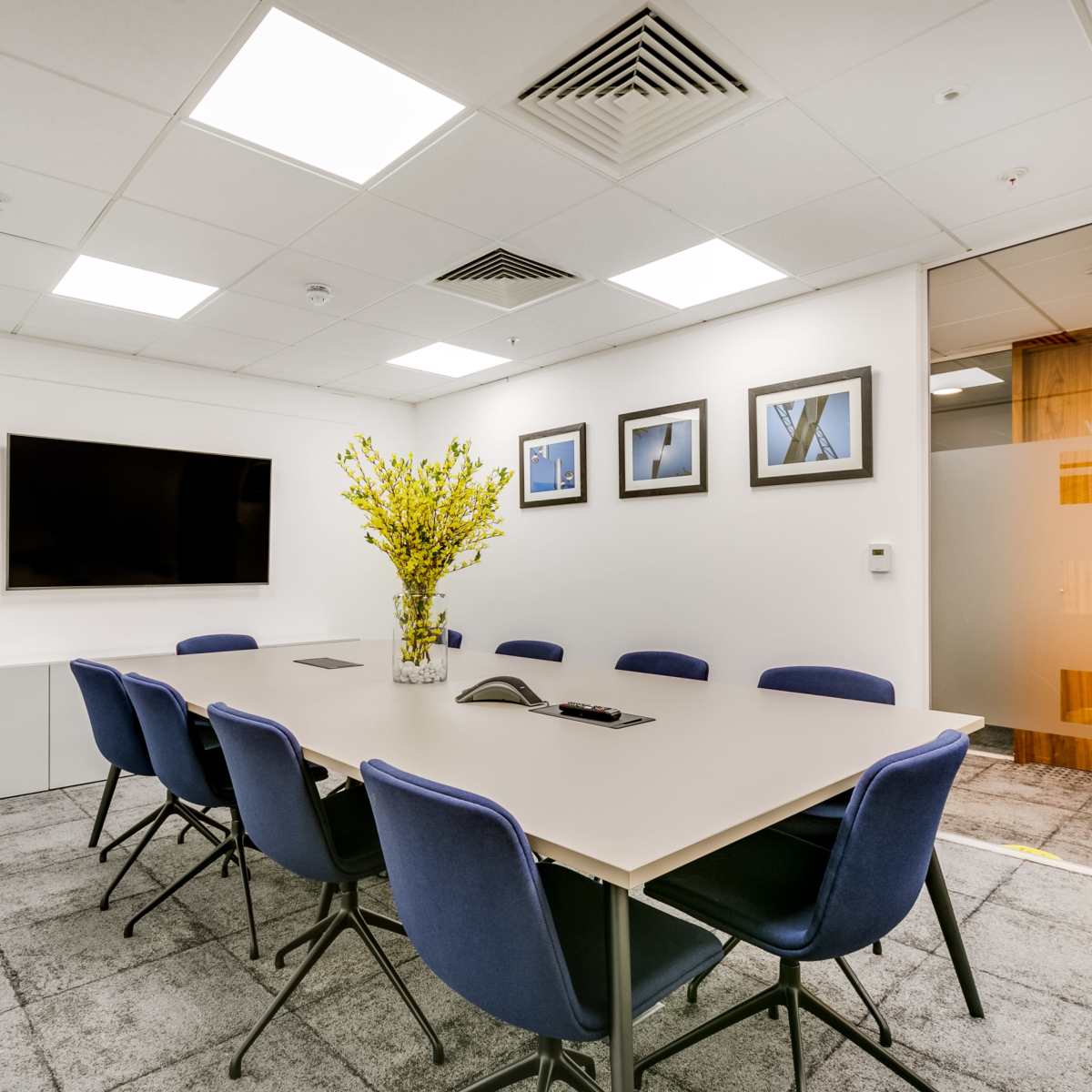 A modern conference room features a long table surrounded by blue chairs, a television on the wall, and a large flower arrangement on the table.