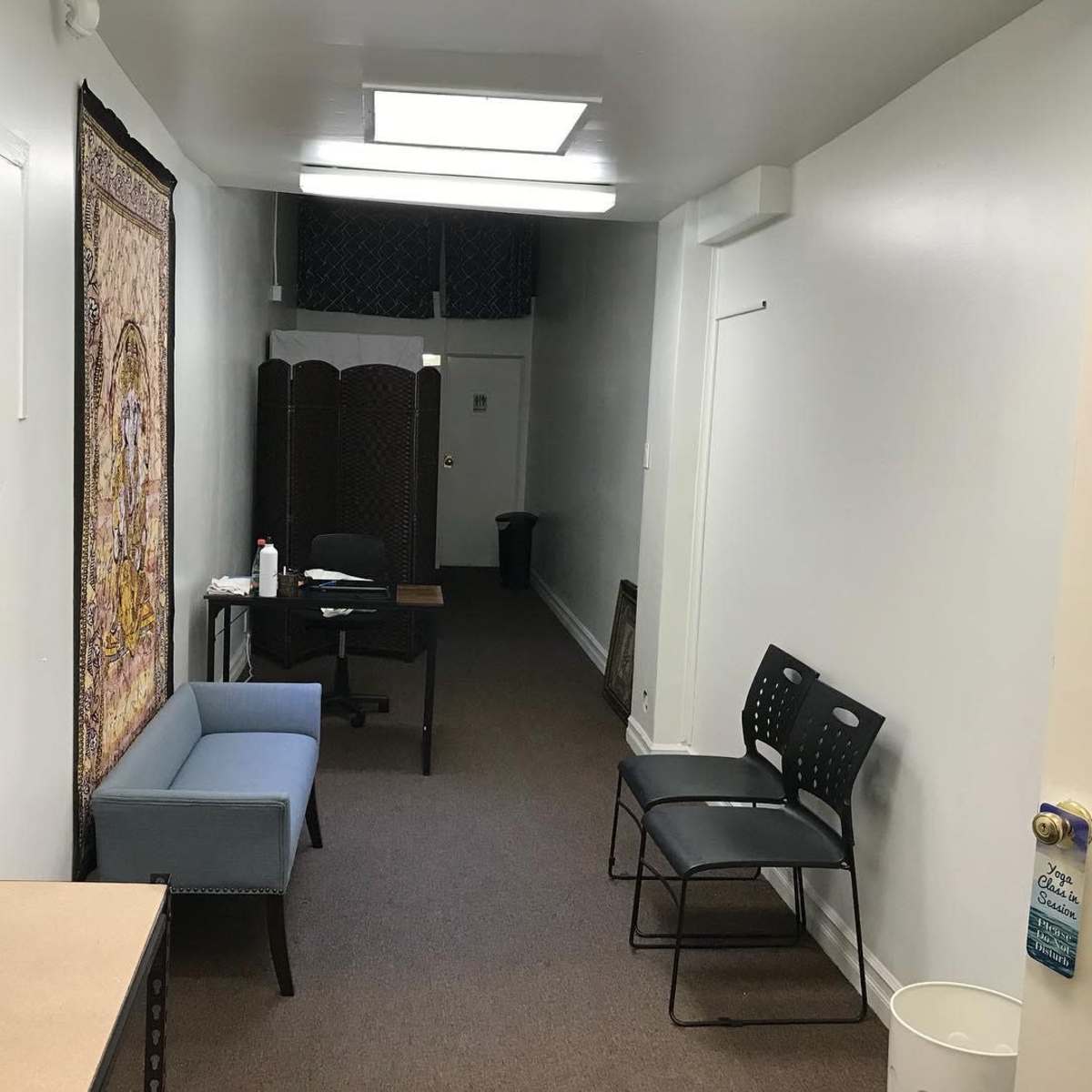 A narrow hallway with a carpeted floor, featuring a small table with a chair, two chairs against the wall, a decorative tapestry, and a wastebasket at the end.